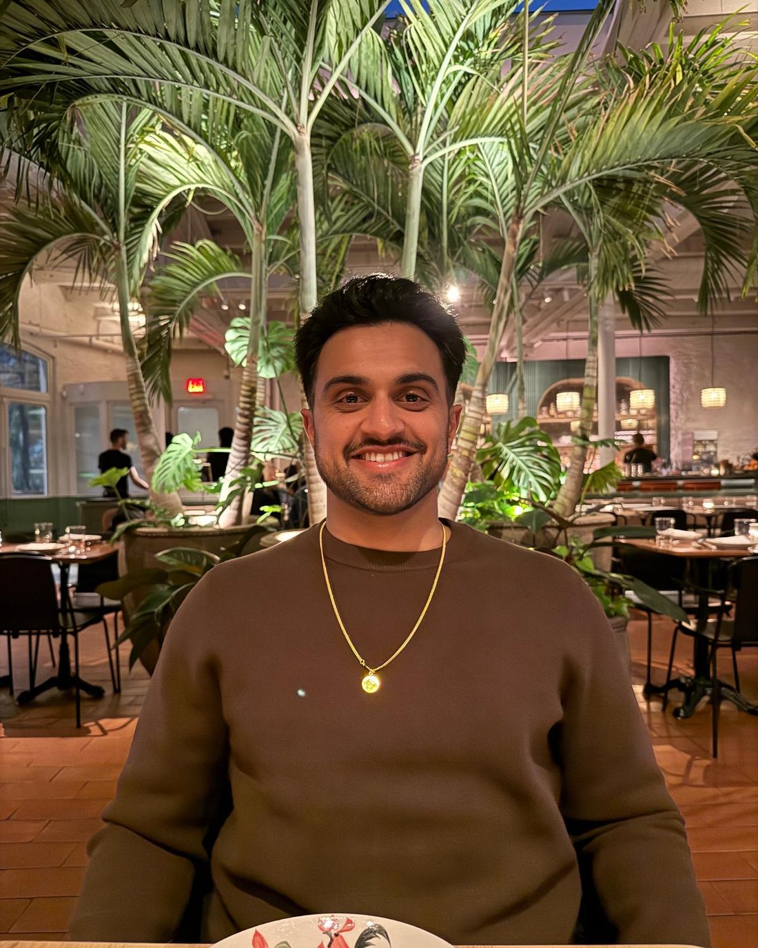 Kiran Aiyar smiling in a warm, plant-filled restaurant setting.