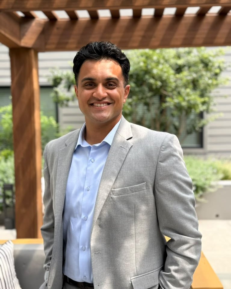 Kiran Aiyar smiling outdoors under a wooden pergola, warm daylight and greenery behind him.