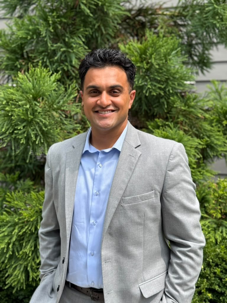 Kiran Aiyar standing outdoors in front of greenery.